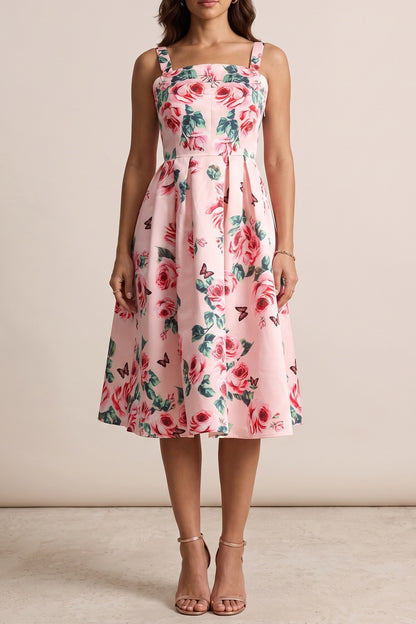 A woman modeling a sleeveless pink midi dress with a red and green floral print, featuring a fitted bodice and a flared skirt, set against a plain beige studio background.