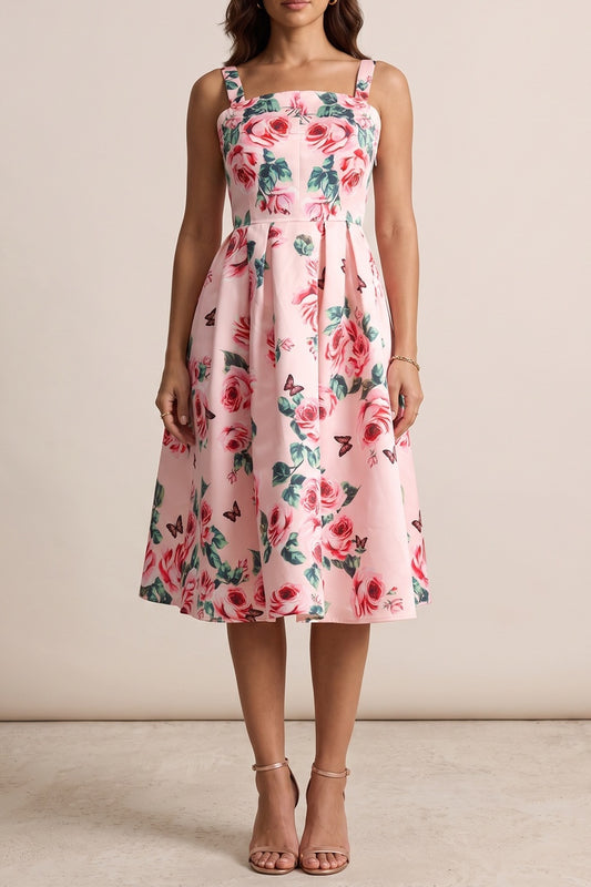 A woman modeling a sleeveless pink midi dress with a red and green floral print, featuring a fitted bodice and a flared skirt, set against a plain beige studio background.