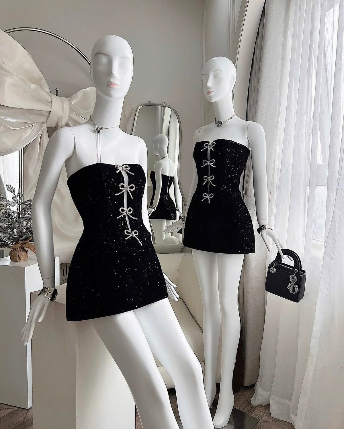 Two mannequins wearing black dresses with white bow details in a room with white curtains.