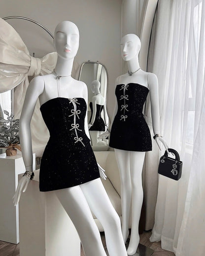 Two mannequins wearing black dresses with white bow details in a room with white curtains.