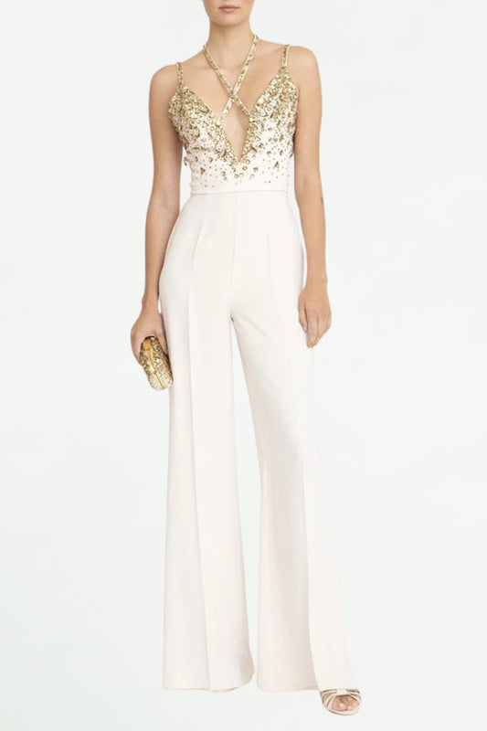 A model wearing a V-neck sequin halter jumpsuit in white, with a floral belt buckle, holding a clutch, and wearing heels.
