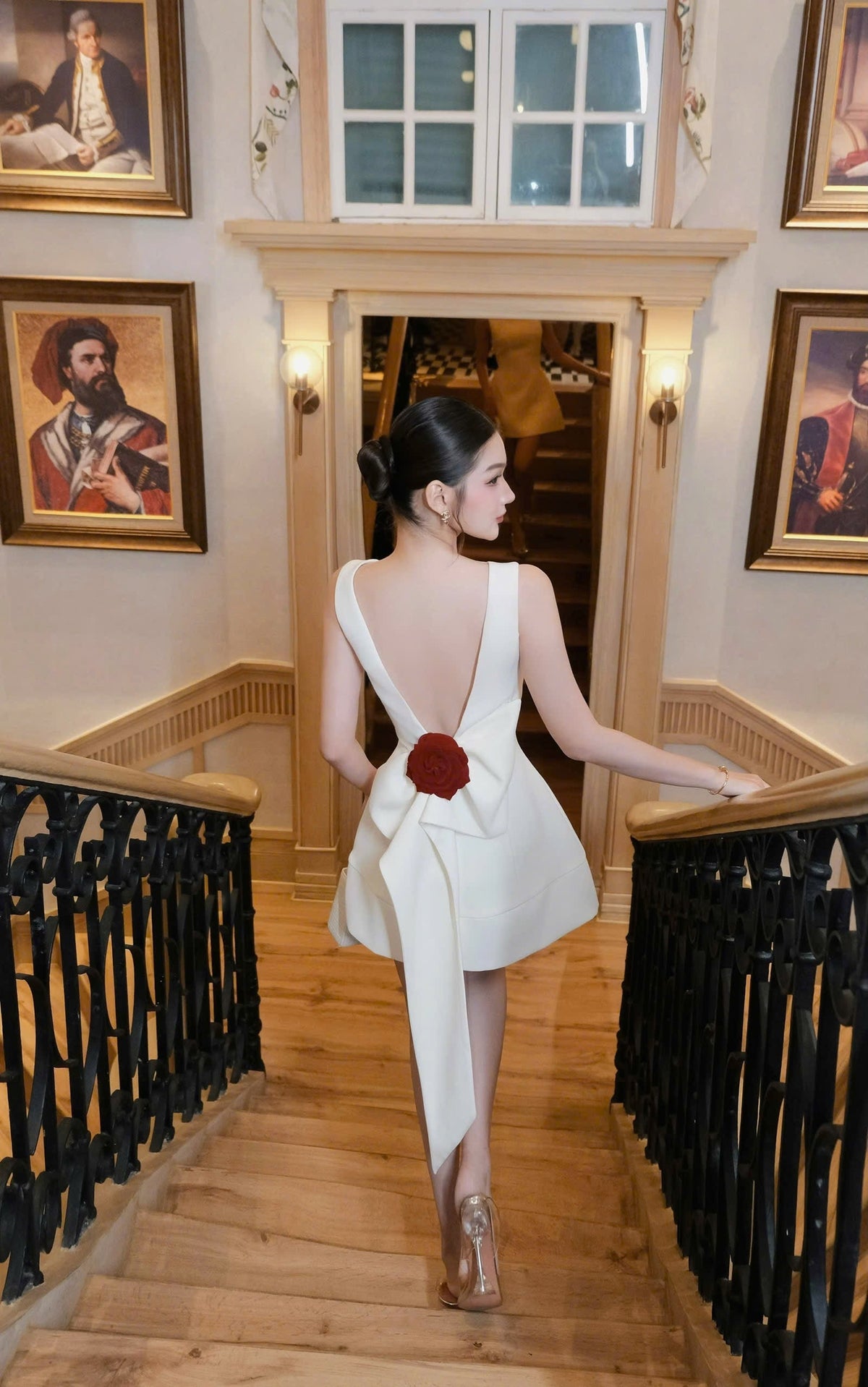 Woman in a white dress with a red flower walking down a staircase in an elegant interior setting.