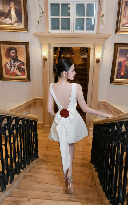 Woman in a white dress with a red flower walking down a staircase in an elegant interior setting.